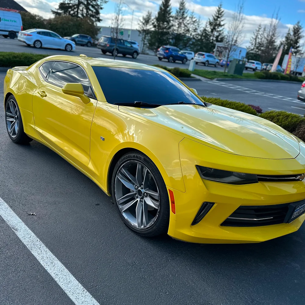 Bright yellow Chevrolet Camaro with deep black window tint parked in a lot under cloudy sky reflections