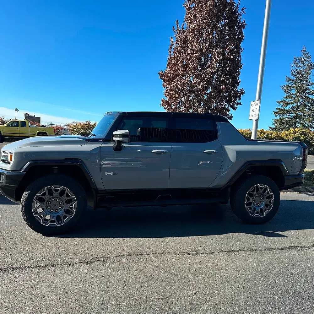 Gray GMC Hummer EV with dark tinted windows and black roof parked in bright daylight near trees
