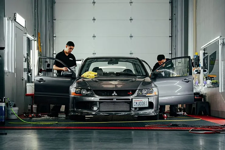 Two detailing technicians working on a gray Mitsubishi sedan inside a professional garage, preparing the vehicle for ceramic coating removal or reapplication at a Tacoma-based auto care facility.