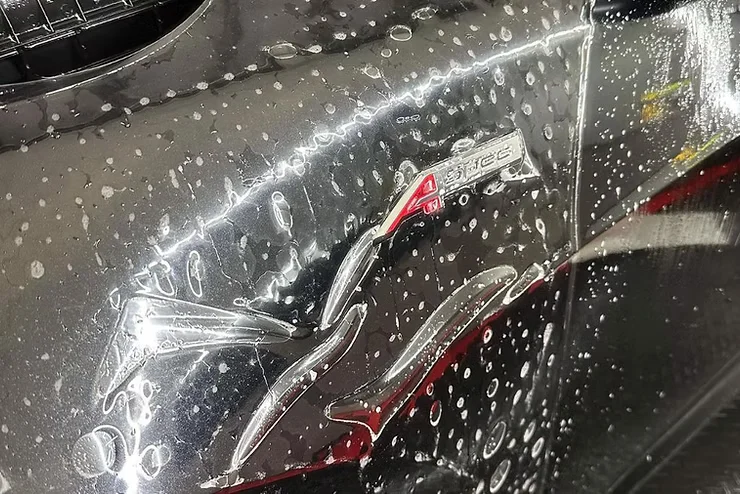 Close-up of a wet car panel with paint protection film freshly applied over minor scratches near a badge labeled “A-Spec,” showing how PPF can conceal surface imperfections on a glossy black finish.