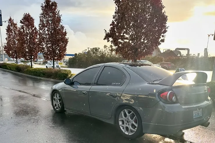 A gray sedan parked on a wet Tacoma street after rainfall, with water spots and road grime visible on the body, illustrating how damp weather and urban debris make scratch protection like PPF especially important in the Pacific Northwest.