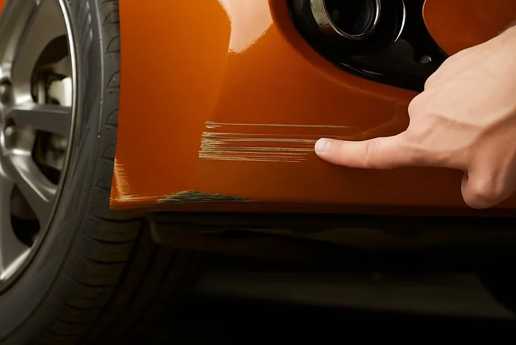 Close-up of a finger pointing to deep scratches on the lower side panel of an orange vehicle, highlighting damage that paint protection film cannot conceal or repair.