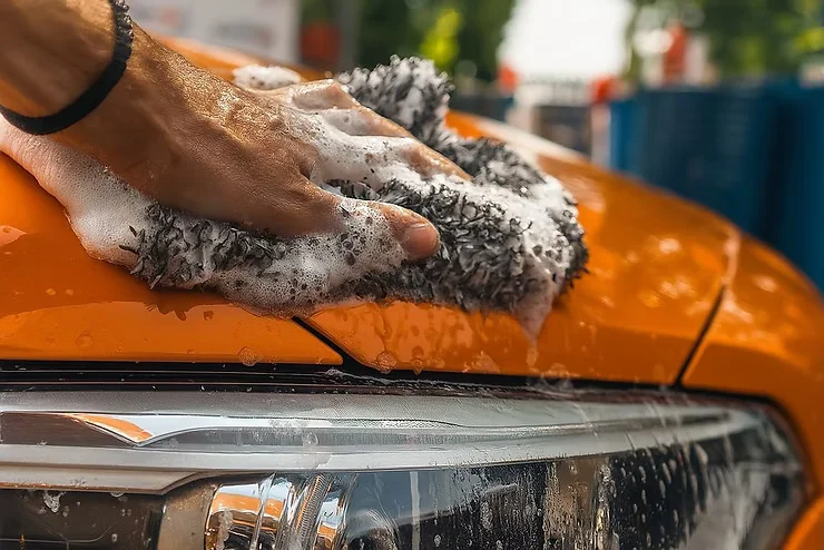 Close-up of a hand washing an orange car hood with a soapy wash mitt, showing how gentle hand washing helps preserve paint protection film and maintain scratch resistance over time.