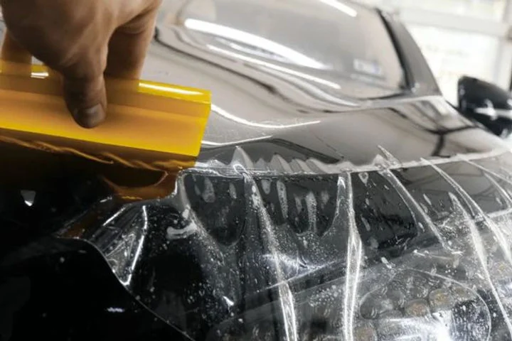 Close-up of a person applying a transparent paint protection film to the hood of a black car using a yellow squeegee, demonstrating how PPF is installed to shield against rock chips and surface damage.
