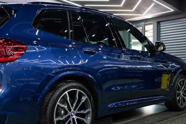 Glossy blue SUV inside a well-lit garage, showing the sleek finish and like-new appearance maintained by paint protection film, with a section of yellow film still visible during the application process.