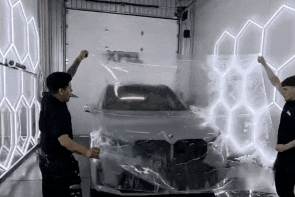 Two technicians stretching clear paint protection film across the front of a black BMW inside a high-tech detailing bay
