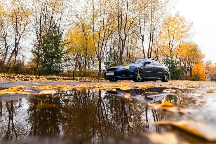 Dark blue car parked on a wet road surrounded by fallen autumn leaves and golden trees, with a large rain puddle reflecting the vehicle and sky