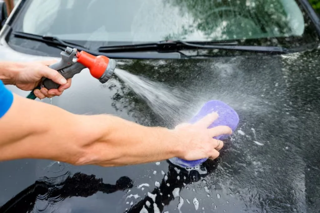 Person hand-washing a black car with a sponge and hose spray to maintain Paint Protection Film (PPF)