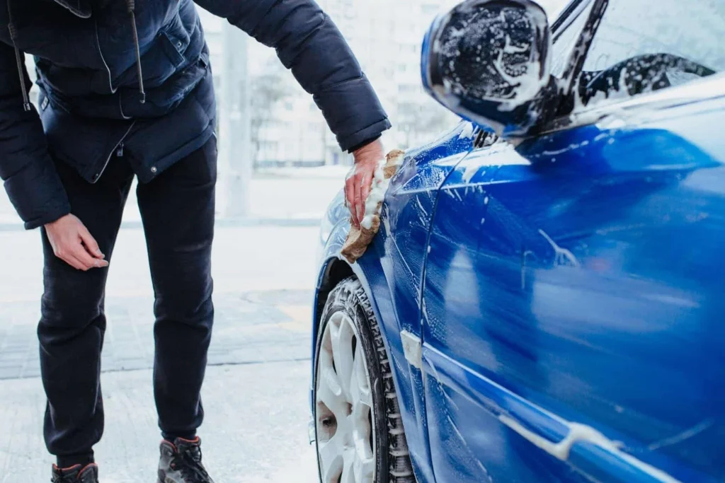 Person washing a blue vinyl-wrapped car using the two-bucket method with a microfiber mitt, focusing on the wheel arch.