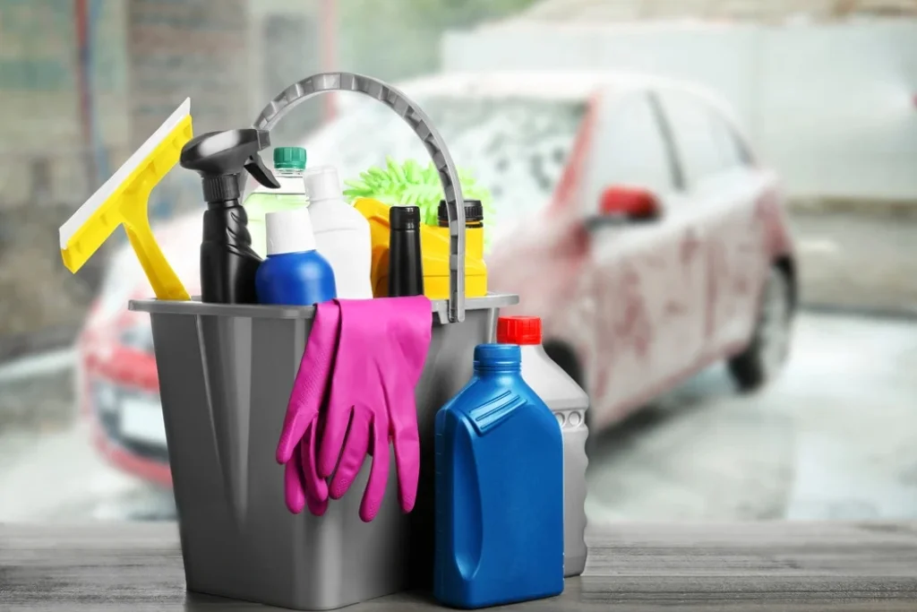 A bucket filled with cleaning supplies including gloves, spray bottles, microfiber towels, and a squeegee, with a vinyl-wrapped car being washed in the background.