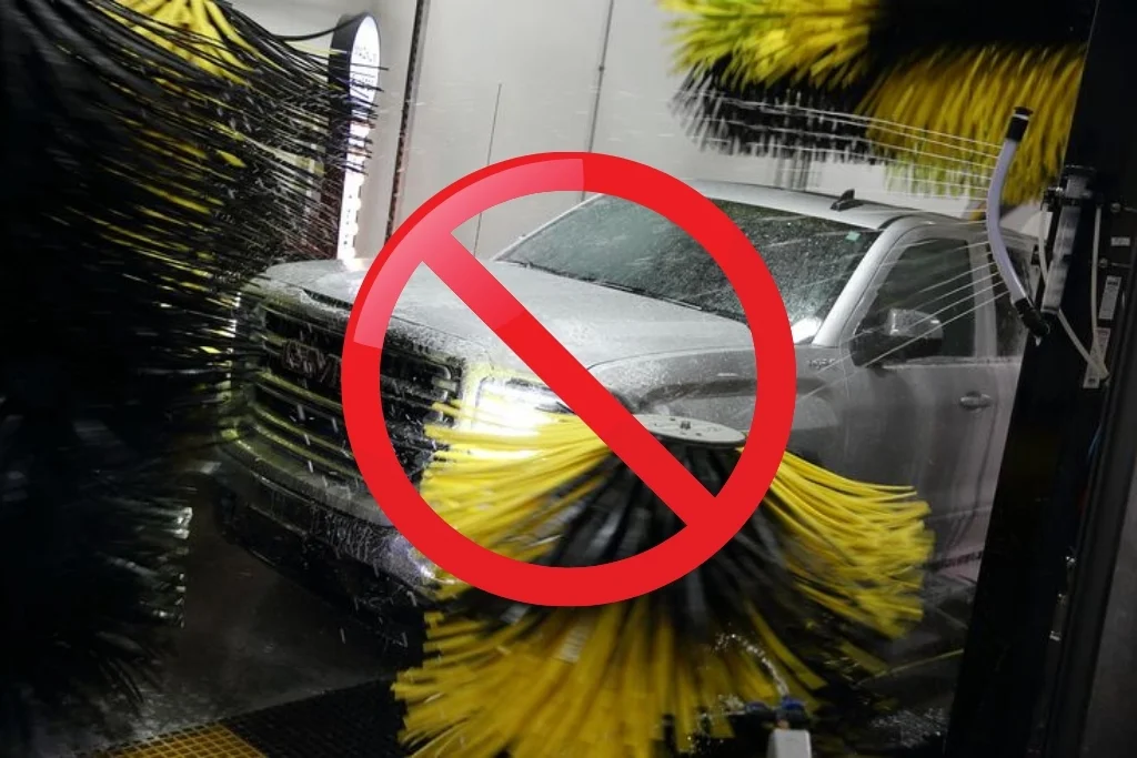 Vehicle going through an automatic car wash with spinning brushes making contact, overlaid with a red prohibition symbol. The image highlights how brush-style automatic washes can scratch or lift vinyl wraps.