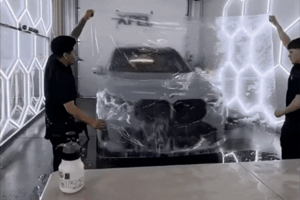 Two technicians installing paint protection film on a silver BMW, applying the clear layer to the car’s hood in a well-lit shop, highlighting PPF’s role in protecting against environmental damage.
