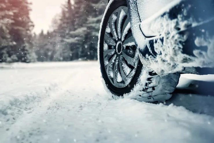 Close-up of a car tire driving through snow, with salt and slush buildup on the vehicle’s lower panels, illustrating winter road hazards.