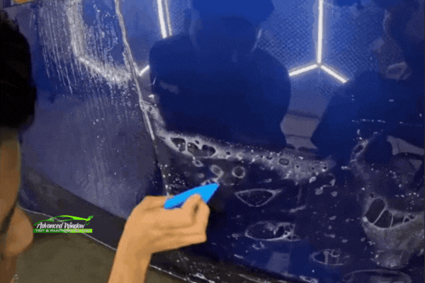 Technician installing paint protection film on a blue car panel in a well-lit shop, demonstrating professional PPF application for winter protection.