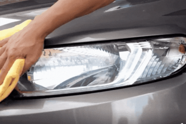 Close-up of a person hand-cleaning a car's headlight and front panel with a yellow microfiber cloth.