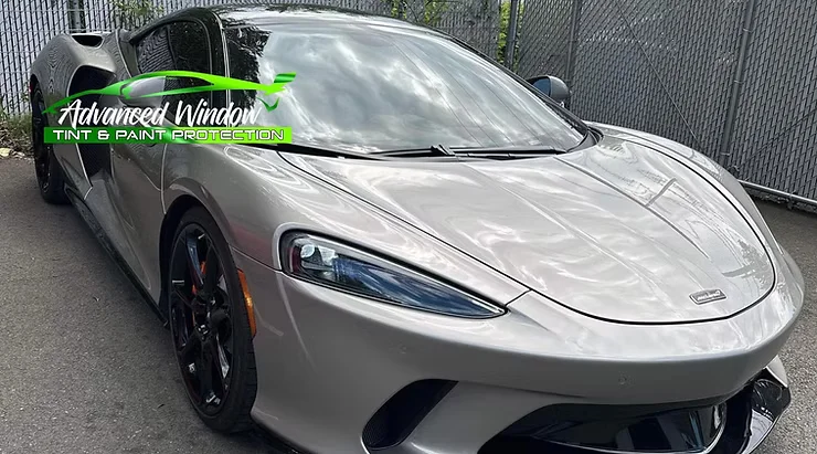 Luxury silver sports car with tinted windows and a high-gloss paint protection film finish, parked near a chain-link fence.