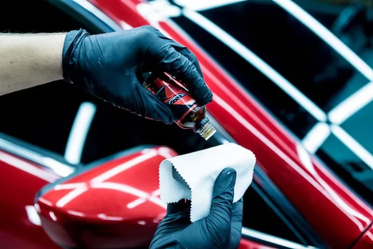 Technician wearing gloves applying ceramic coating to a red vehicle's paint protection film (PPF) using a cloth and applicator bottle.