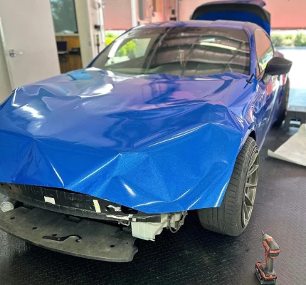 Blue vinyl wrap being applied to front end of sports car with bumper removed inside wrap studio