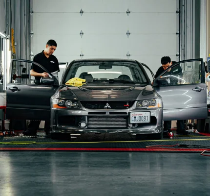 Two technicians applying window tint to a gray Mitsubishi Lancer Evolution with both front doors open in a shop bay