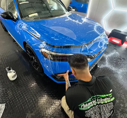 Installer from Advanced Window Tinting applying clear paint protection film to front bumper of blue car in detailing bay