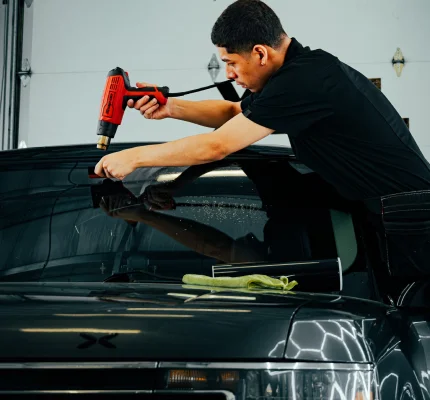 Technician using heat gun to apply window tint film to windshield of black SUV in detailing shop