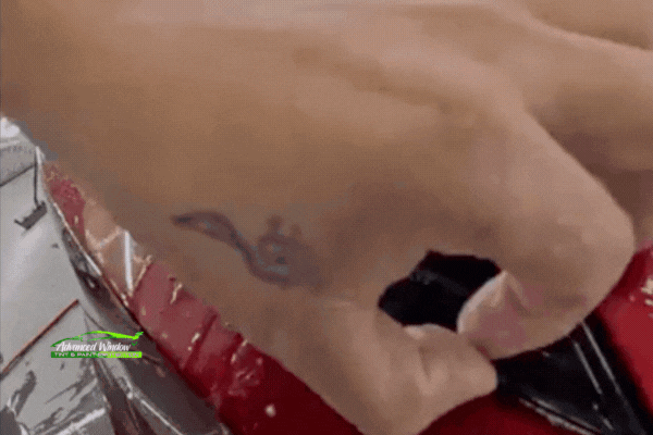 Close-up of a professional applying paint protection film to a headlight on a red vehicle, showing the precision and clean fit of expert installation, with the Advanced Window Tint & Paint Protection logo in the