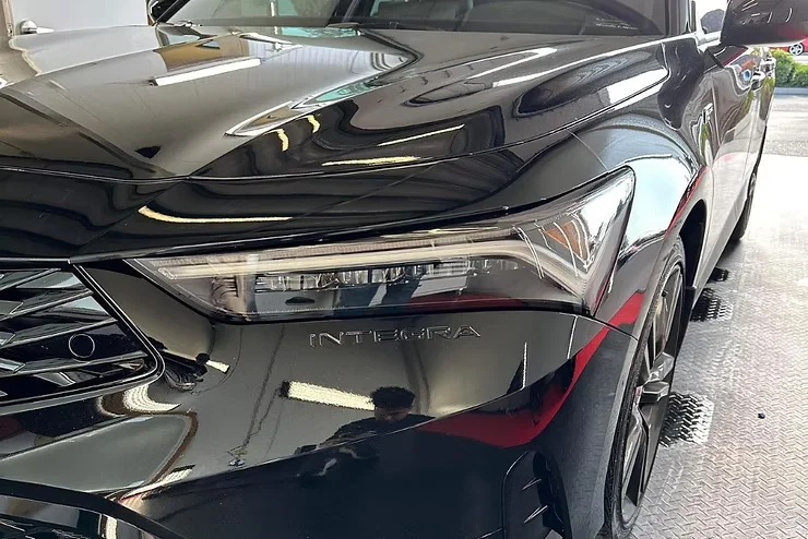 Glossy black Acura Integra in a detailing bay with a protected headlight, reflecting clear overhead lights — illustrating the long-term clarity and durability of professionally installed paint protection film.