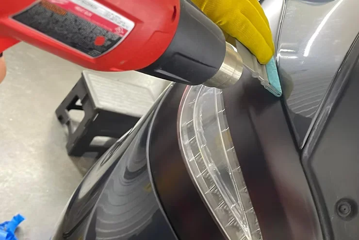 Technician using a heat gun and gloved hand to apply paint protection film to a car’s headlight and adjacent body panel, demonstrating the precise installation process and protection benefits.