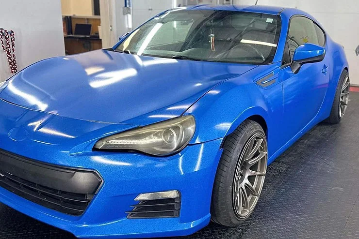 Bright blue vinyl-wrapped sports car with a satin finish parked inside a detailing shop, showcasing a completed custom wrap