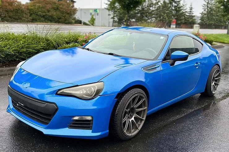Glossy blue vinyl-wrapped sports car parked on wet pavement with greenery and trees in the background, highlighting the finished wrap in natural light
