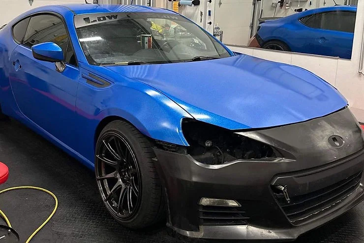 Blue vinyl-wrapped sports car with an unfinished matte black front bumper in a shop, showing the flexible and reversible nature of vehicle wrapping