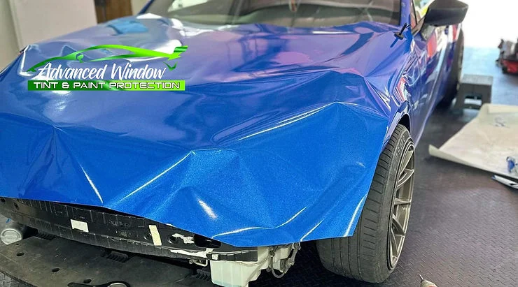 Blue vinyl wrap partially applied to a sports car in a professional shop, with the front bumper removed during installation