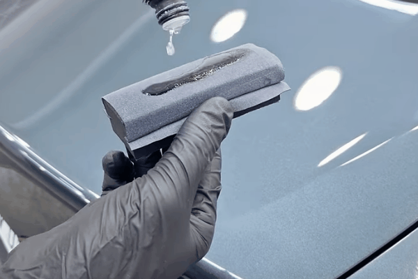 A gloved hand holds a foam applicator pad while ceramic coating liquid is dripped onto it, preparing for application on a glossy car surface. The image illustrates the beginning step of bonding ceramic coating to a vehicle’s paint.
