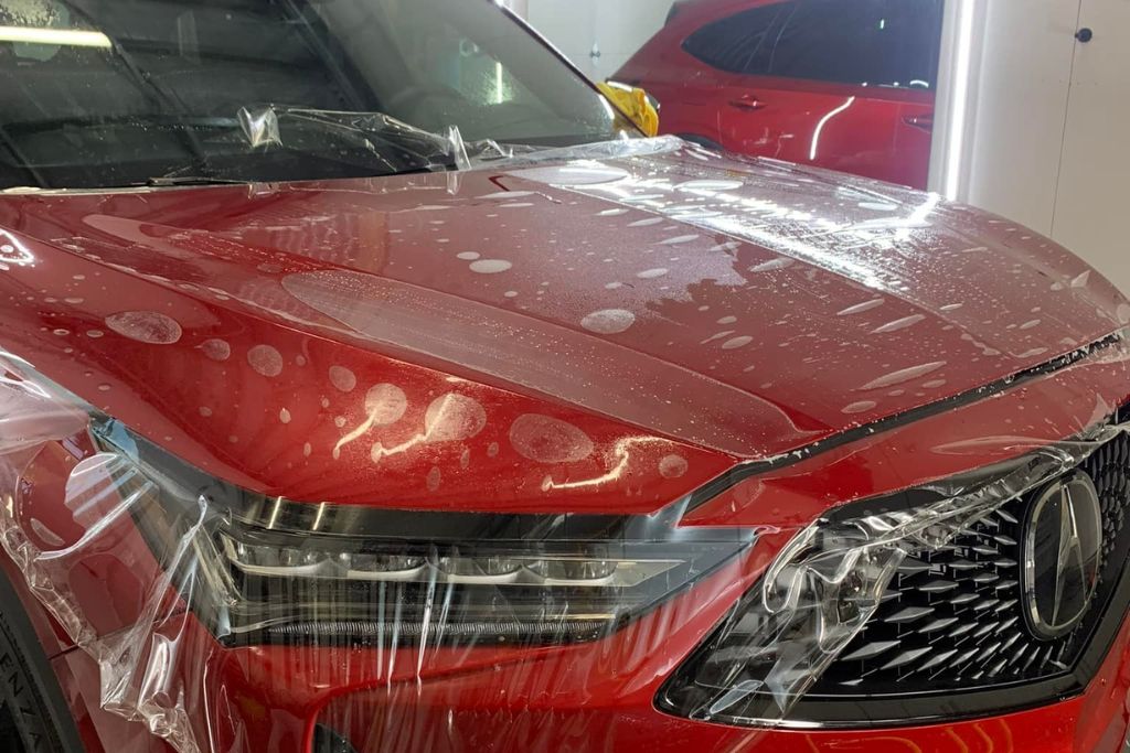Close-up of a red SUV with paint protection film being installed on the hood and fenders. The film is partially applied, with visible air bubbles and moisture on the surface, highlighting the early stage of the PPF installation process.