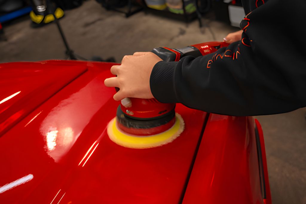 Person using a dual-action polisher with a yellow foam pad to correct the paint on a red vehicle, an essential step before reapplying ceramic coating.