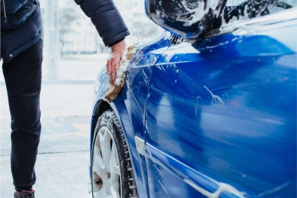 Person hand-washing a blue car with a soapy mitt, focusing on the front fender and wheel area, demonstrating safe maintenance practices for ceramic-coated vehicles.