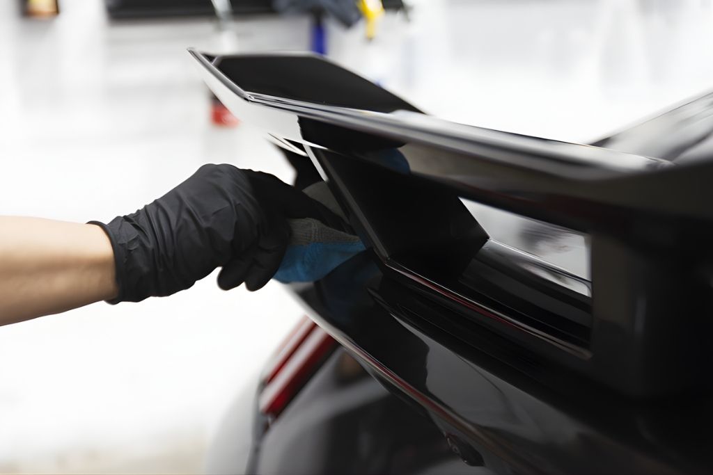 A person wearing a black glove applies ceramic coating to a glossy black car spoiler using a microfiber cloth, demonstrating precision detailing during reapplication.