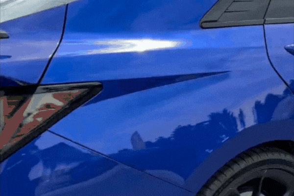 Close-up of a highly reflective blue car panel showing the deep gloss and mirror-like finish achieved through a ceramic coating, with surrounding sky and trees clearly reflected on the surface.