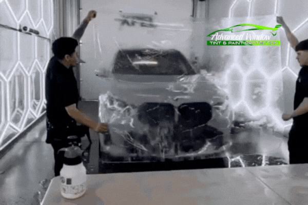 Two technicians apply a large sheet of clear paint protection film to the front of a dark-colored BMW inside a well-lit detailing bay. The process showcases Advanced Window Tinting & PPF’s precision installation using XPEL film.