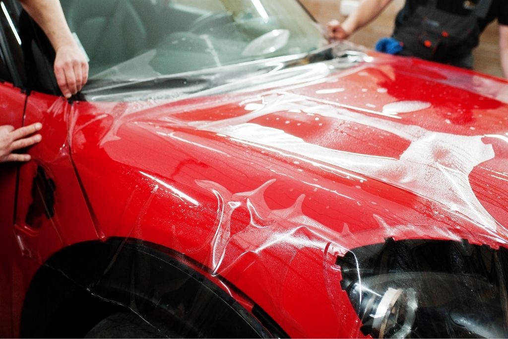 Technicians installing paint protection film on a red car hood during professional PPF application.