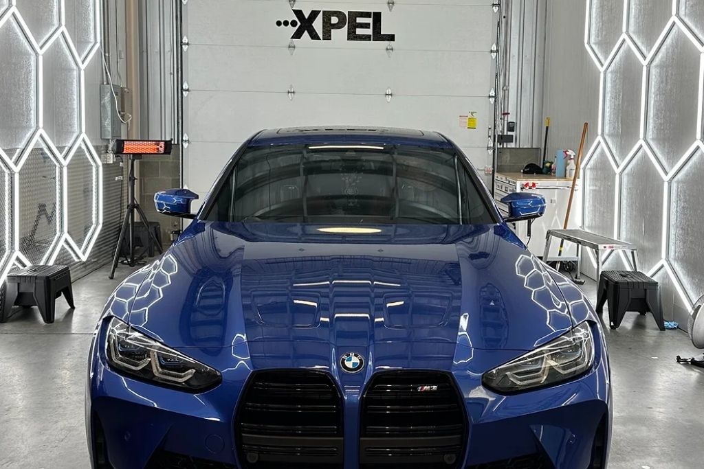 Front view of a blue BMW M3 parked inside a professional detailing studio with XPEL branding visible in the background.