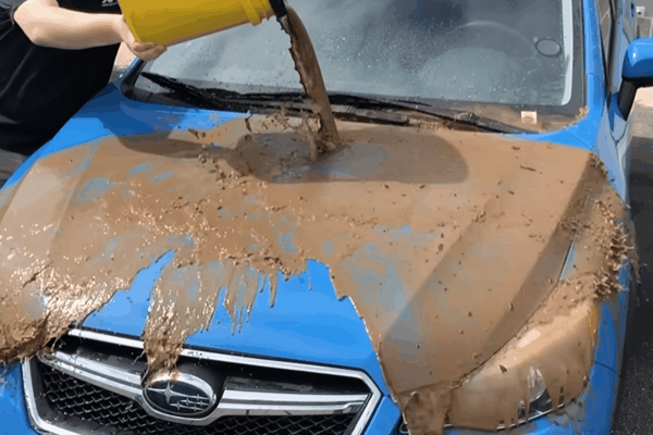 A person pours a bucket of muddy water over the hood of a blue Subaru, showing how the liquid slides off cleanly without sticking. Demonstrates the hydrophobic, self-cleaning effect of ceramic coating under dirty, wet conditions.