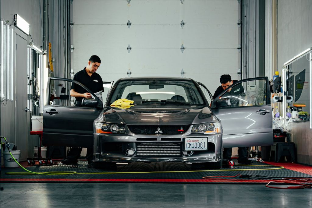 Two technicians work on a silver Mitsubishi sedan inside a well-lit detailing shop, with both front doors open and supplies visible nearby. The car has a Washington license plate, emphasizing the importance of professional ceramic coating application in wet climates.