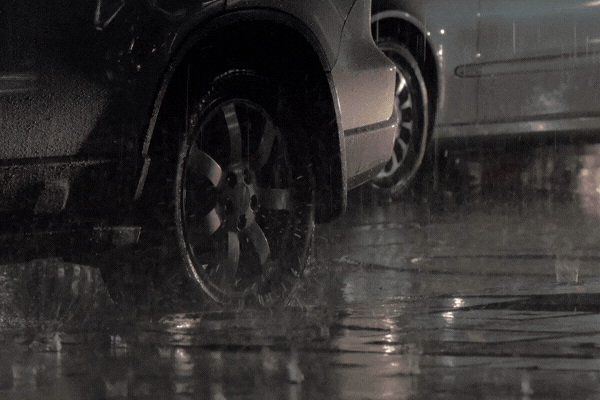 Close-up of two parked cars during heavy rain at night, with water splashing around the tires and reflective puddles forming on the ground. Highlights how prolonged exposure to wet conditions can lead to paint damage and corrosion.