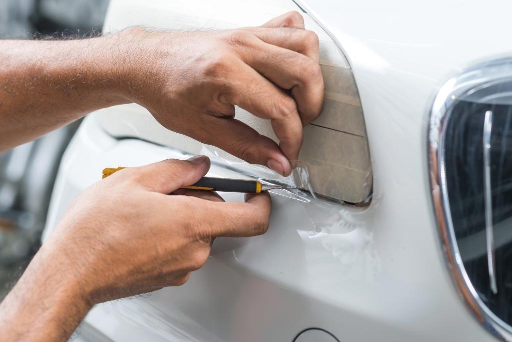 Technician trimming and applying paint protection film around a car headlight during professional PPF installation.