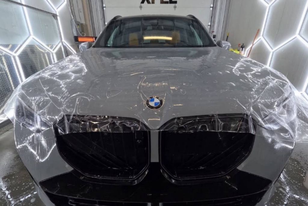 Paint protection film being installed across the hood and front end of a gray BMW in a professional detailing bay.