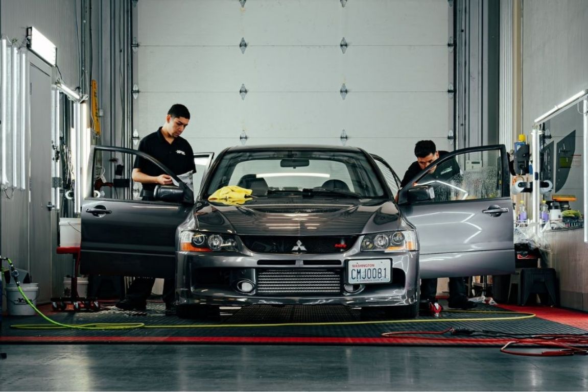 Two automotive technicians working on a gray Mitsubishi inside a professional detailing and vehicle protection shop.