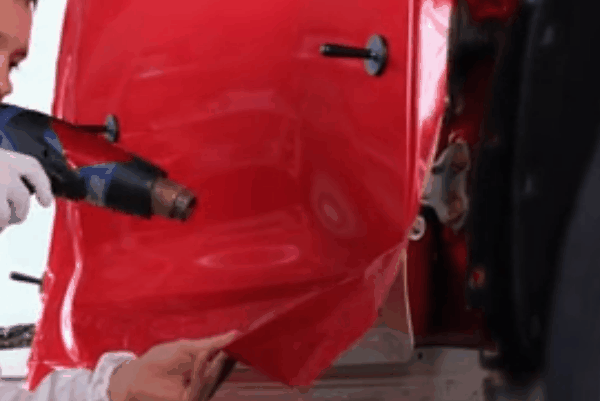 Technician heating and stretching red vinyl wrap over a vehicle body panel during installation.