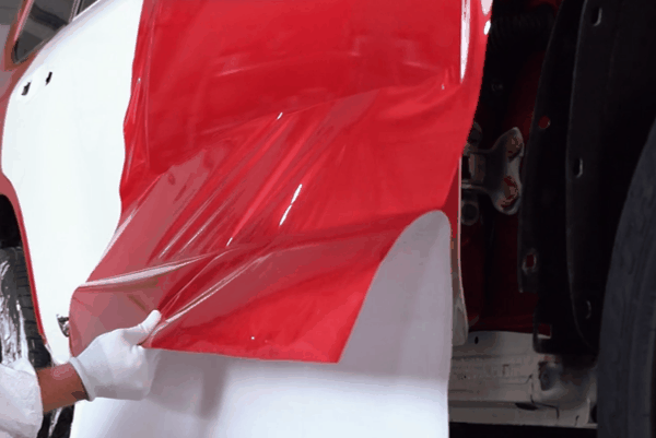 Technician heating and stretching red vinyl wrap over a vehicle body panel during installation.