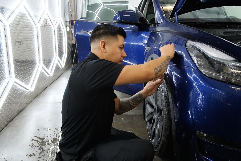 Technician installing paint protection film on the front fender of a blue vehicle in a professional detailing shop.